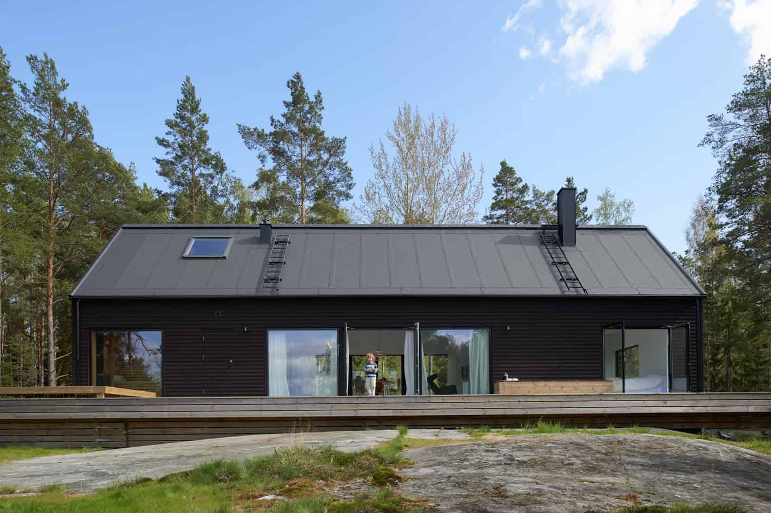 Contemporary black wooden house with large glass sliding doors, located in a lush forest setting under a blue sky with clouds, showcasing modern architecture and design.