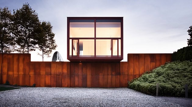 Contemporary modern house with sleek glass facade and rust-colored metal exterior, surrounded by minimalist landscaping and a gravel driveway, showcasing innovative architecture and stylish design.