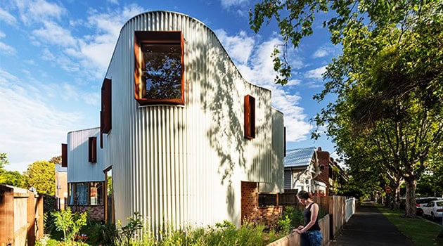 Futuristic modern house with curved metal exterior and large windows surrounded by greenery and trees in a suburban neighborhood.