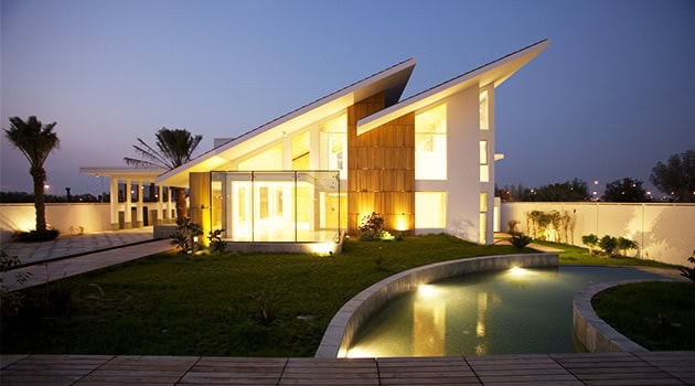 Sleek modern house with futuristic architectural design, illuminated at night, featuring sharp angular rooflines and large glass windows in a tranquil landscaped setting.
