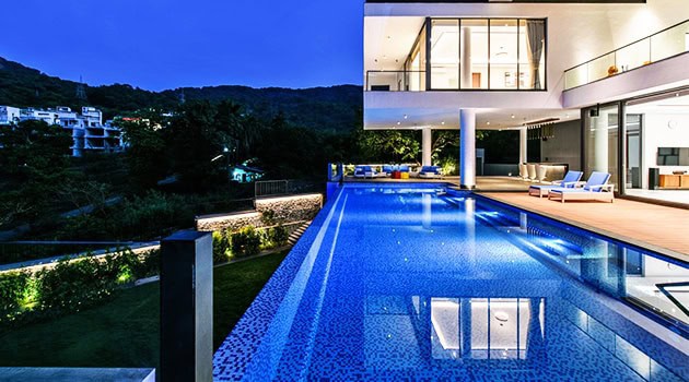 Luxurious modern house with illuminated infinity pool and contemporary architecture, overlooking scenic mountains and lush greenery at dusk.