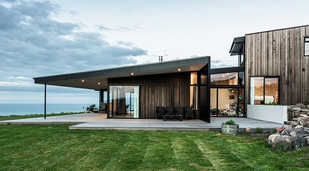 Contemporary modern house with large glass windows, wooden exterior, and expansive outdoor patio overlooking scenic landscape and ocean views.