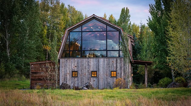 Modern rustic wooden house with large glass windows and a unique geometric roof set in a lush forest landscape.