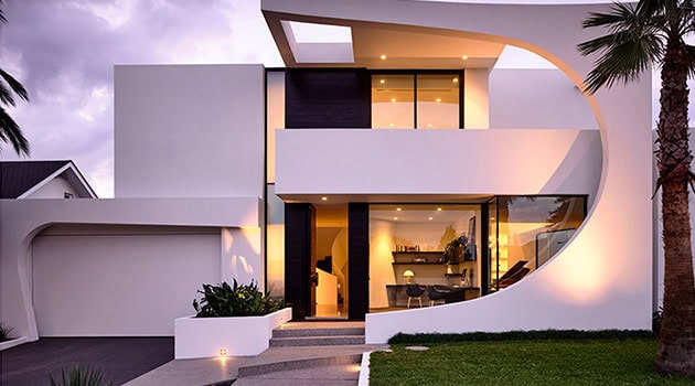 Modern contemporary white house with curved architectural design and large glass windows, surrounded by palm trees and landscaped yard.