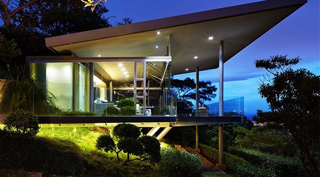 Sleek modern house with glass walls and overhanging roof, set in a lush landscape during evening with exterior lighting.