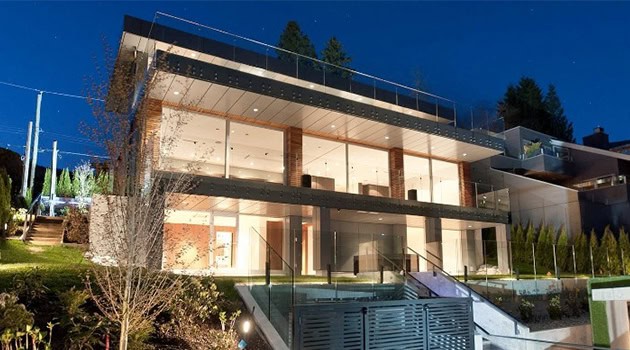 Modern luxury glass house with multiple levels, expansive outdoor terraces, and sleek architectural design nestled in a lush landscape at night.