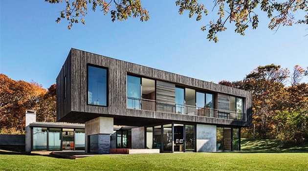 Contemporary modern house with sleek wooden and concrete exterior, large glass windows, and spacious balcony overlooking a lush green landscape.
