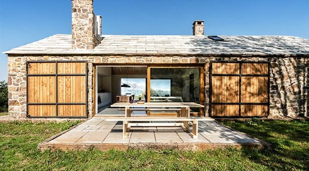Rustic stone and wood cottage exterior with large folding doors opening to outdoor patio and scenic ocean view, showcasing traditional architecture and modern design elements.