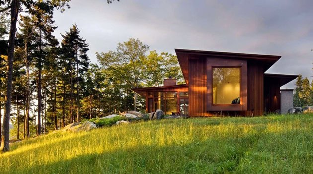 Modern wooden house with angular roof and large windows set in lush green landscape during sunset.