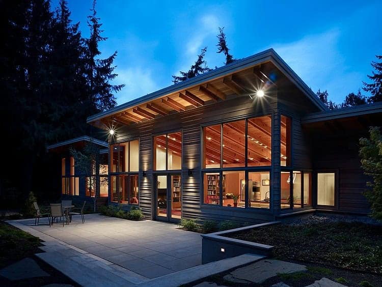 Modern wooden house with large windows and outdoor patio at dusk, showcasing contemporary architecture and design.