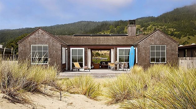 Modern beach house with wooden exterior, large windows, and outdoor deck, set against dunes and coastal landscape, perfect for beachfront living and serenegetaway retreats.