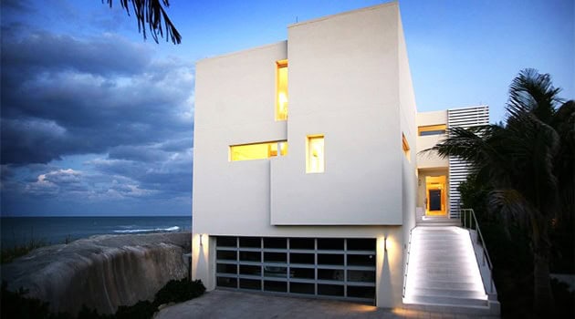 Modern minimalist beachfront house with clean geometric lines and large windows, set against a dramatic evening sky, showcasing innovative architectural design and oceanfront living.