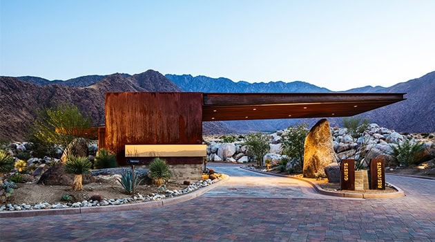 Modern desert residential house with contemporary architecture, rust-colored steel facade, and desert landscaping, set against mountain backdrops.