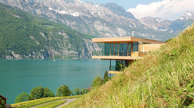 Modern lakeside glass house with sleek architecture overlooking a serene turquoise lake and majestic mountains in the background.