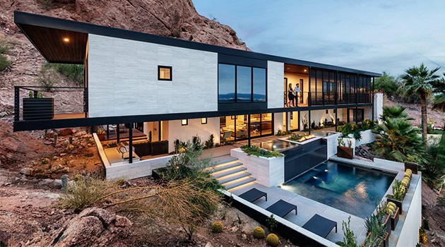Luxury modern hillside mansion with sleek architectural design, large glass windows, outdoor pool, and contemporary landscaping, fitting seamlessly into a desert landscape.
