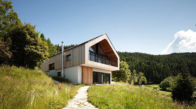 Modern wooden house with large windows and a sloped roof in a lush green landscape, surrounded by forest and nature.