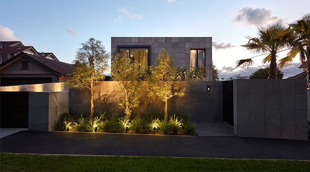 Contemporary modern house with dark stone exterior, lush landscaping, and outdoor lighting, showcasing innovative architecture and stylish design elements.