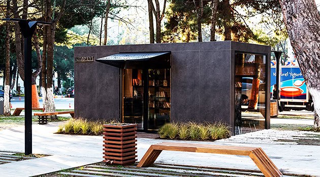 Compact modern black glass and concrete pavilion nestled in a lush park setting, featuring contemporary architectural design with sleek lines and minimalist aesthetic.
