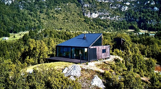 Modern minimalist mountain cabin with large glass windows, located in lush green forest landscape, showcasing innovative architectural design and sustainable building materials.