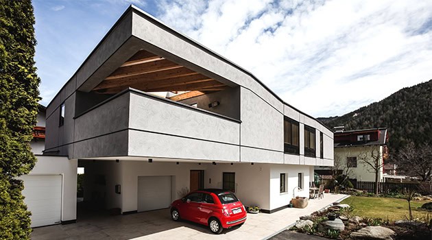 Modern minimalist residential building with sharp lines and a distinctive overhang, featuring concrete exterior and large windows, surrounded by natural landscape.