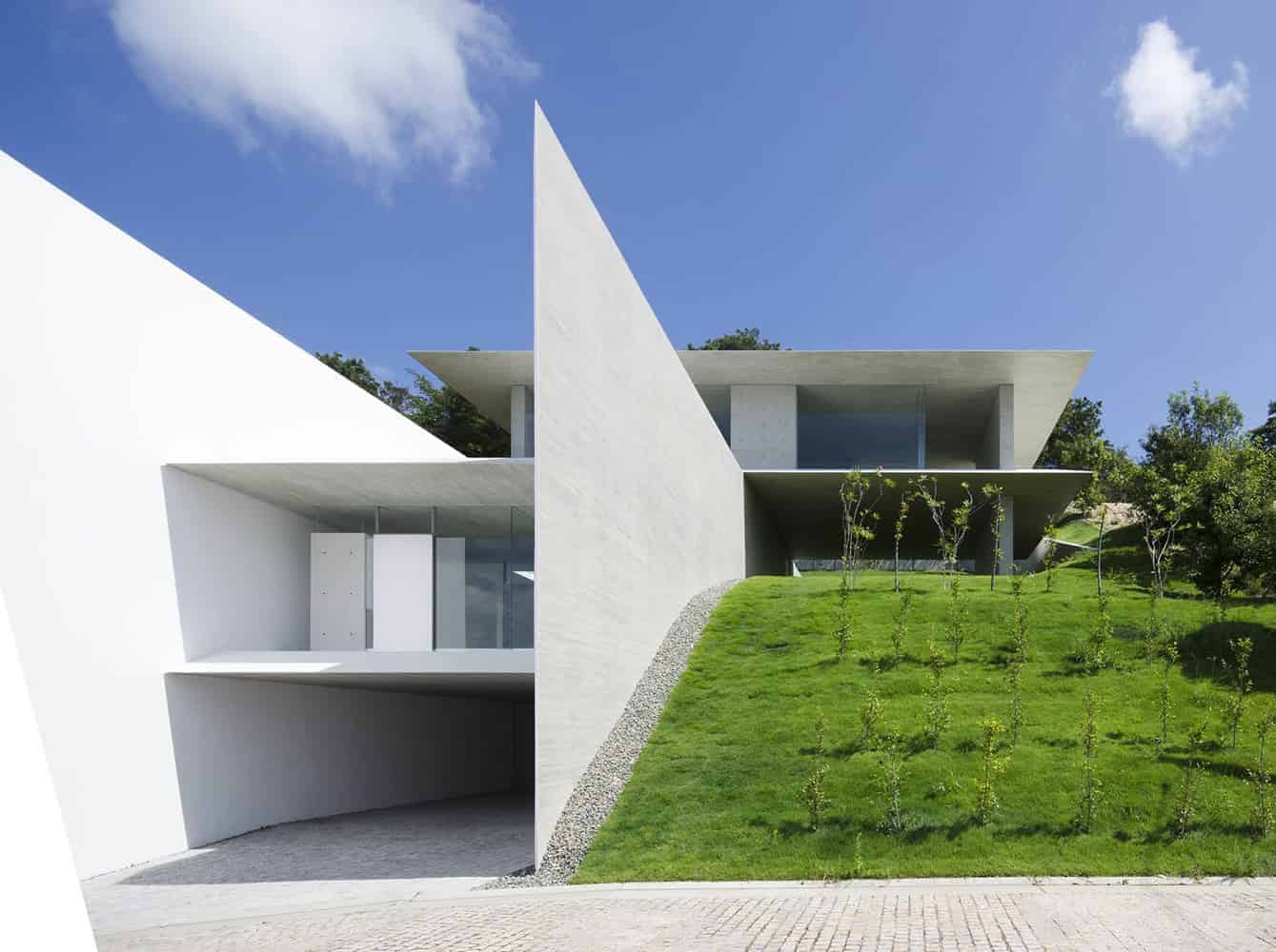 Modern minimalist white concrete house with sharp geometric design and large glass windows, set against a bright blue sky with lush greenery.