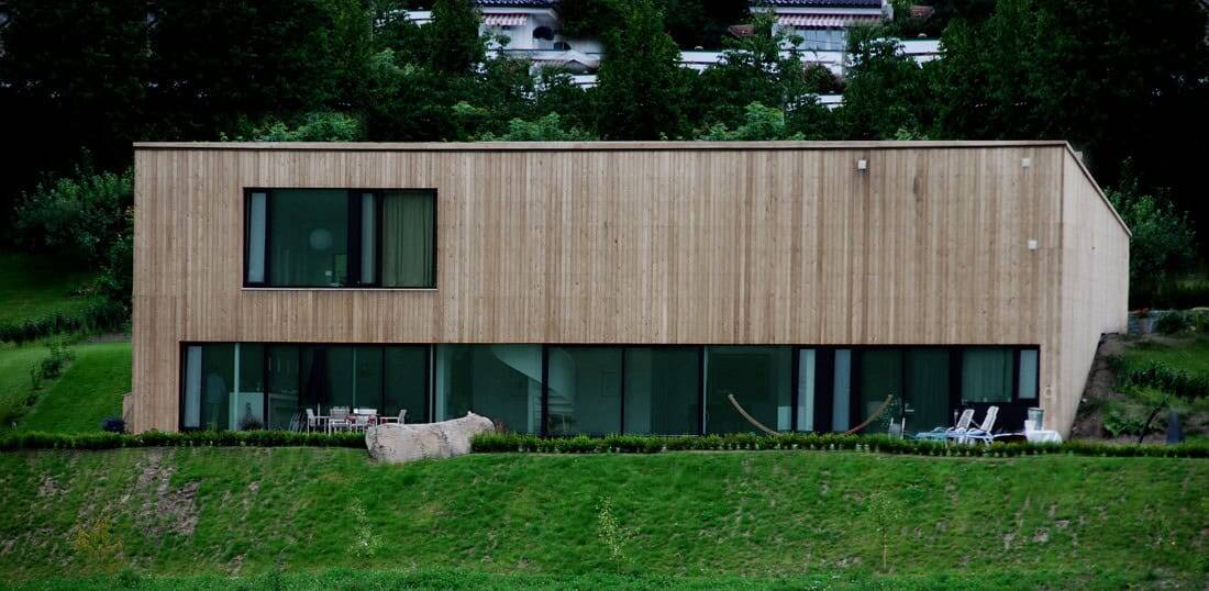 Modern minimalist house with wooden exterior and large glass windows, situated in lush green landscape.