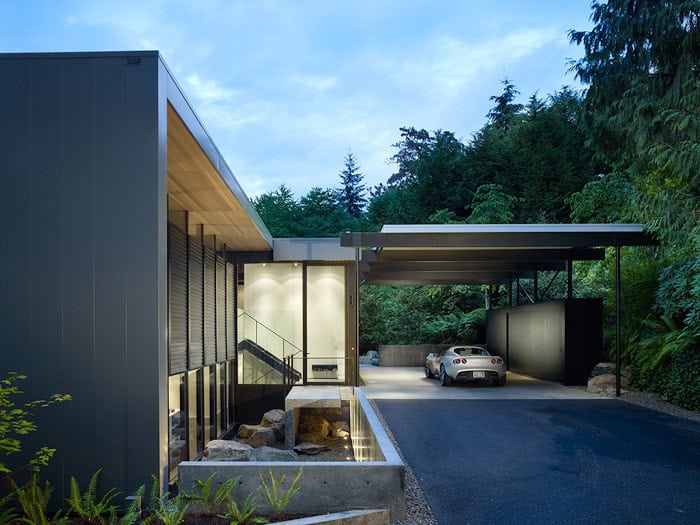 Contemporary modern house with sleek design, large windows, and a spacious carport set against lush green trees.