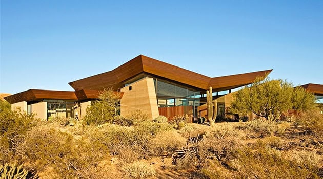 Sleek modern desert house with angular rooflines and extensive glass walls surrounded by arid desert landscape and native plants.