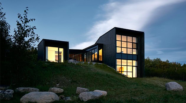 Contemporary modern house with large glass windows, black exterior, and innovative architectural design set on a grassy hillside during dusk.
