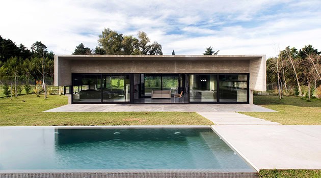 Modern minimalist house with large glass windows and flat roof overlooking a sleek swimming pool in a lush outdoor setting.