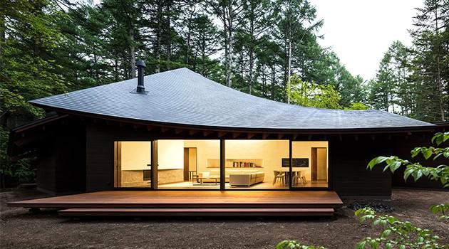 Modern minimalist black wood house with large glass doors showcasing an open-plan interior amidst lush greenery.