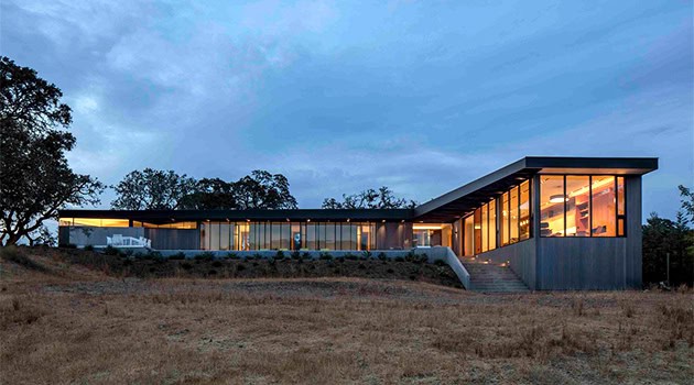 Contemporary modern house with extensive glass windows and sleek design, set on a grassy landscape during twilight.