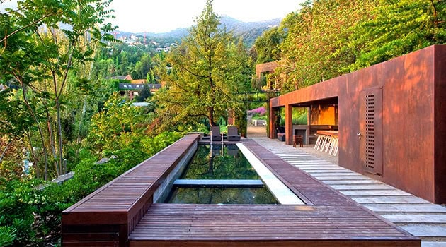 Modern hillside house with sleek wooden deck and rust-colored exterior walls surrounded by lush greenery and mountain views for contemporary architecture and landscape design.