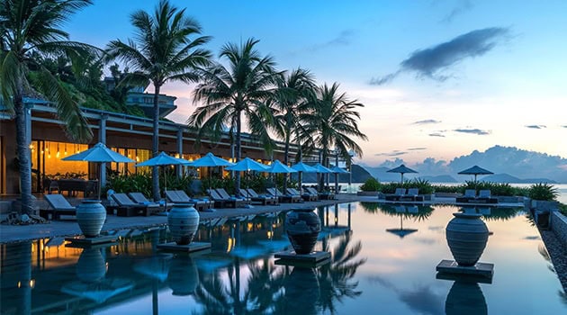 Luxurious tropical resort poolside with palm trees, sun loungers, and umbrellas at sunset, showcasing modern architecture and serene atmosphere.