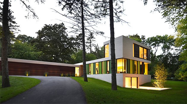 Modern minimalist house with large glass windows and vibrant yellow accents, surrounded by lush greenery and tall trees.