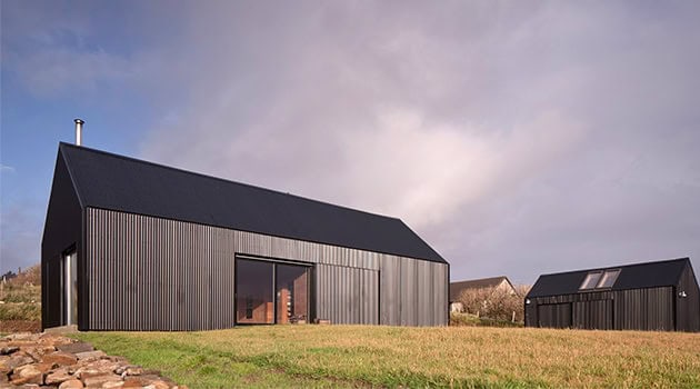 Modern black metal barn-style house with large glass doors and a sloped roof, set in a rural landscape with a cloudy sky.