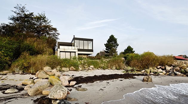 Modern beachside house with large glass windows and minimalist design, surrounded by natural landscape and rocky shoreline, showcasing contemporary architecture and coastal living.