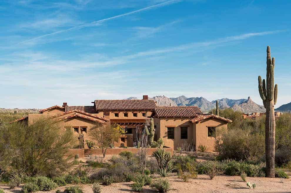 Modern Southwest desert house with adobe-style architecture surrounded by desert plants and cacti in a mountainous landscape during daytime.