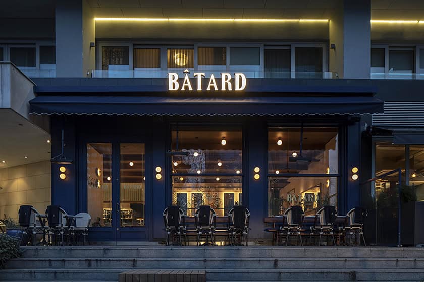 Bâtard restaurant exterior with stylish modern architecture, large glass windows, elegant outdoor seating, and warm ambient lighting, showcasing contemporary urban dining design.