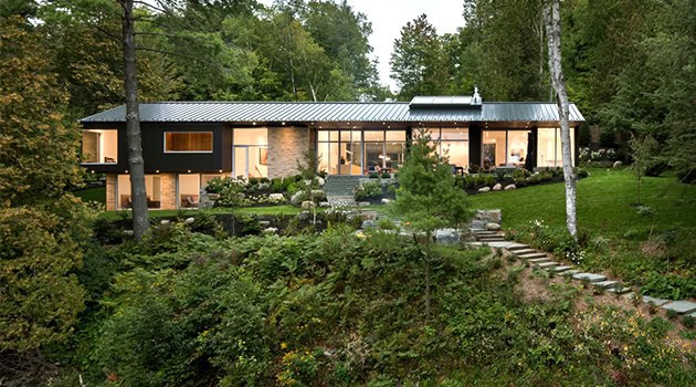 Spacious modern house with large glass windows, surrounded by lush greenery and landscaped gardens in a wooded area.