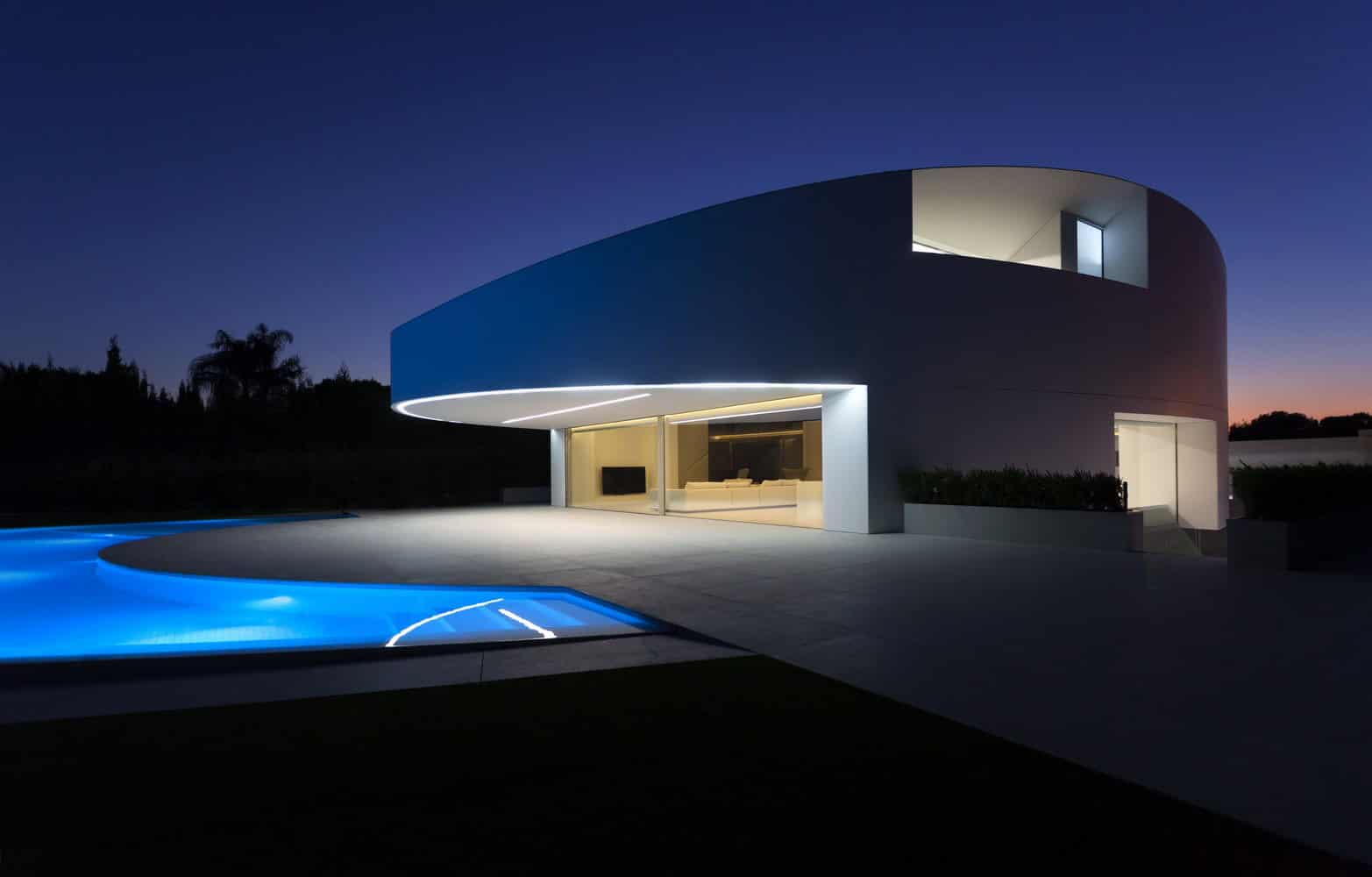 Ultra-modern minimalist house with smooth white exterior, large glass windows, and glowing exterior lighting, featuring a sleek design with a pool under a twilight sky.