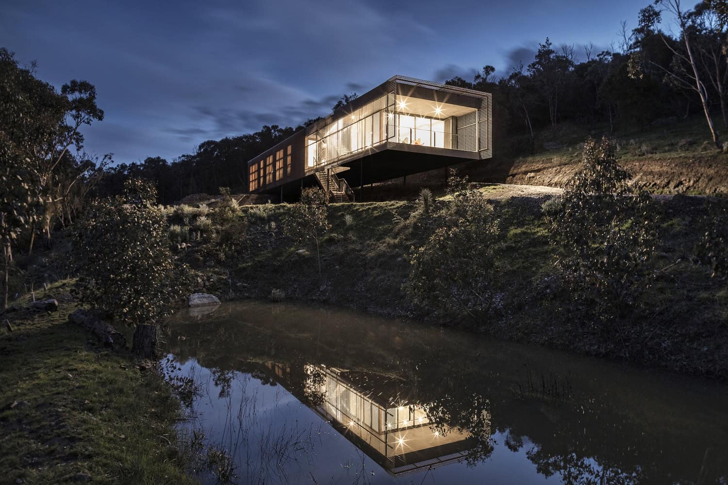 Contemporary modern house on a hillside with large glass windows and illuminated interior, reflecting on a nearby water body at night, surrounded by natural landscape and trees.