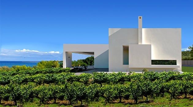 Modern minimalist white house with clean geometric lines overlooking lush greenery and a glimpse of the ocean in the background.