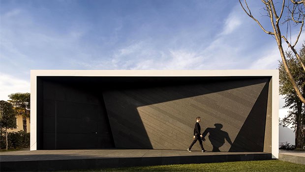 Sleek modern minimalist garage with black textured facade and white frame, featuring geometric design and shadow play, set in a landscaped area with clear blue sky.
