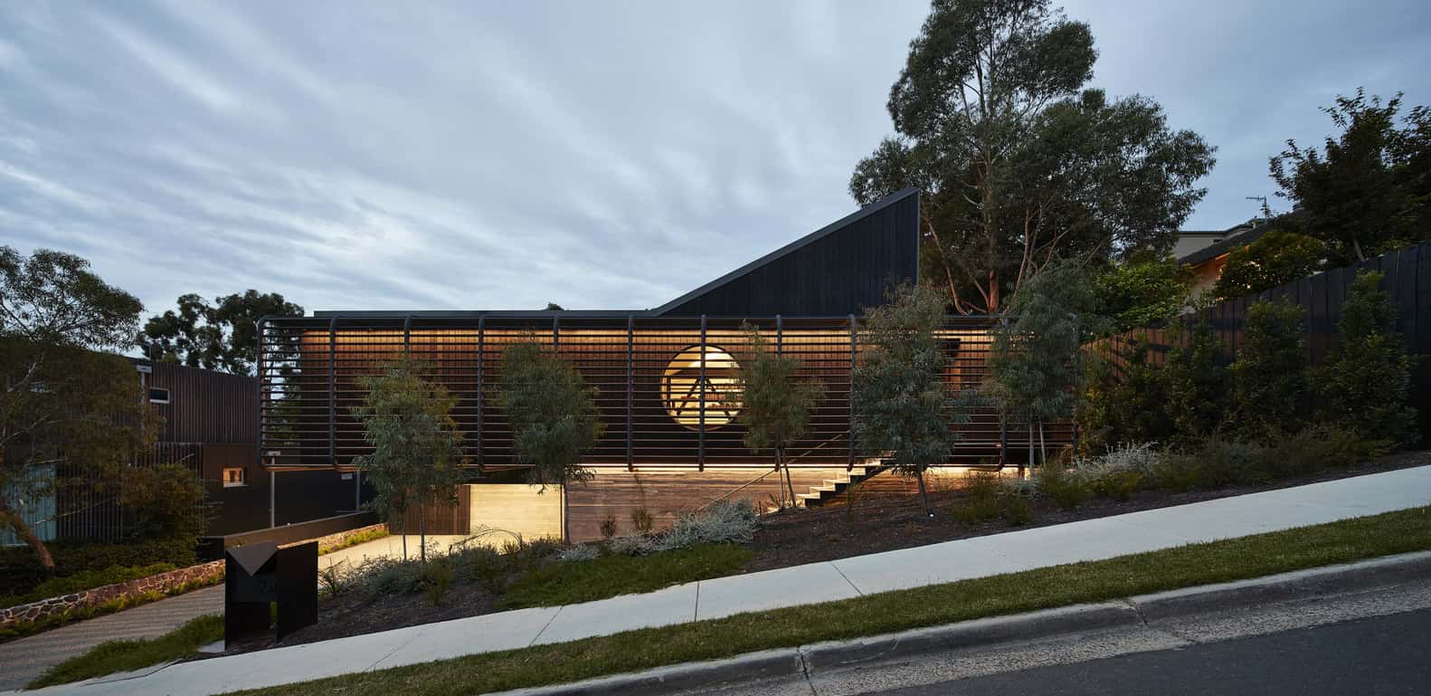 Modern architectural house with sleek black exterior, innovative design, and illuminated circular window, surrounded by lush greenery and landscape lighting.