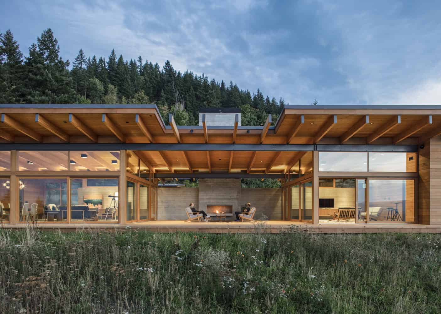 Contemporary modern house with extensive glass walls, wooden accents, and outdoor living space set against a scenic mountain backdrop, embodying innovative architectural design.