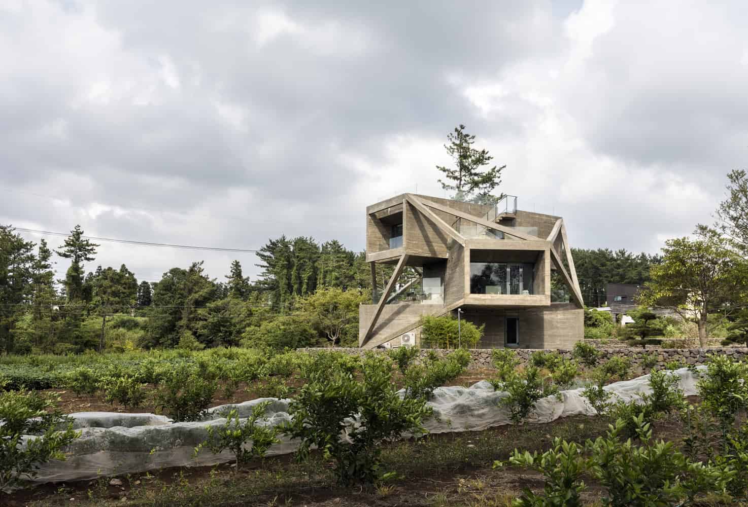 Futuristic modern house with angular concrete architecture and large glass windows, situated amidst lush greenery and farmland under a cloudy sky.