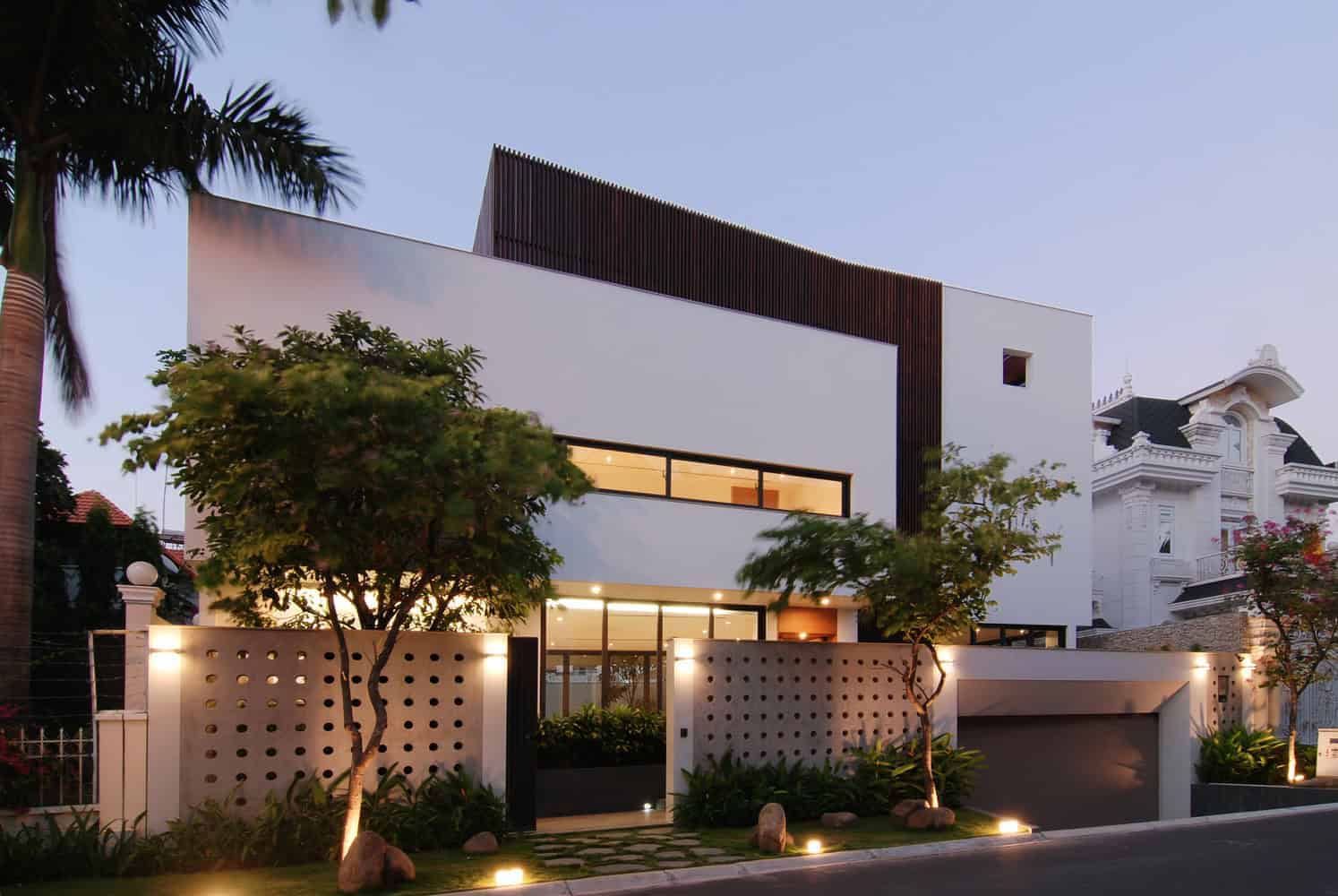 Modern minimalist white house with sleek architectural design, large glass windows, and landscaped outdoor space featuring trees and ambient lighting.