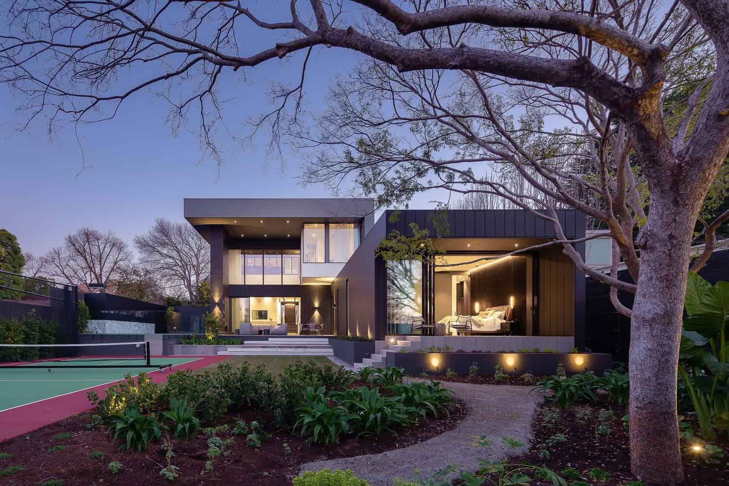 Contemporary modern house exterior with sleek architectural design, large glass windows, and outdoor lighting, surrounded by lush landscaping and mature trees, at dusk.