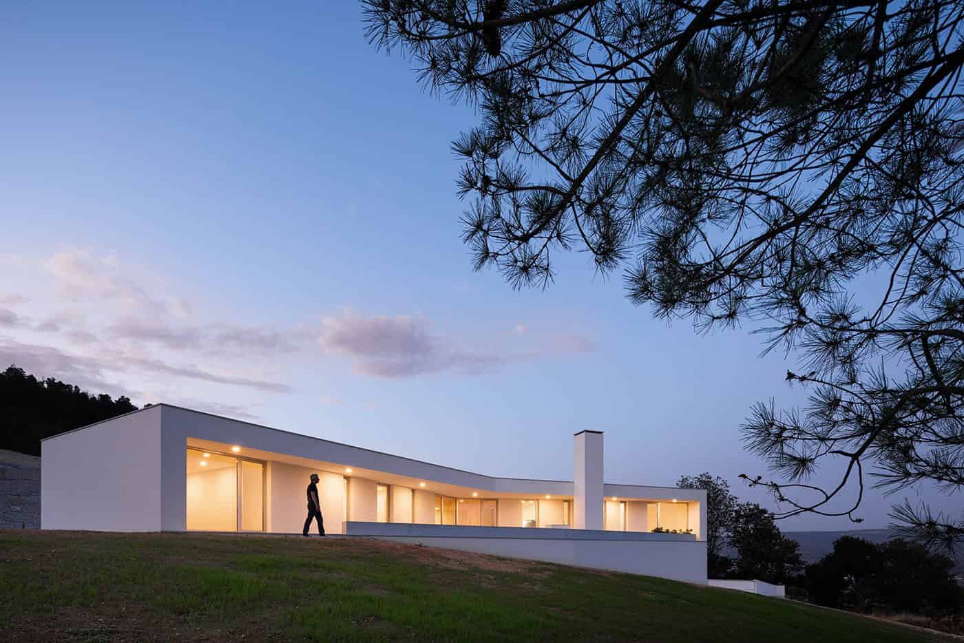Modern minimalist house with sleek white exterior and large glass windows, situated on a grassy hillside during dusk, surrounded by nature, emphasizing contemporary architectural design and outdoor living spaces.